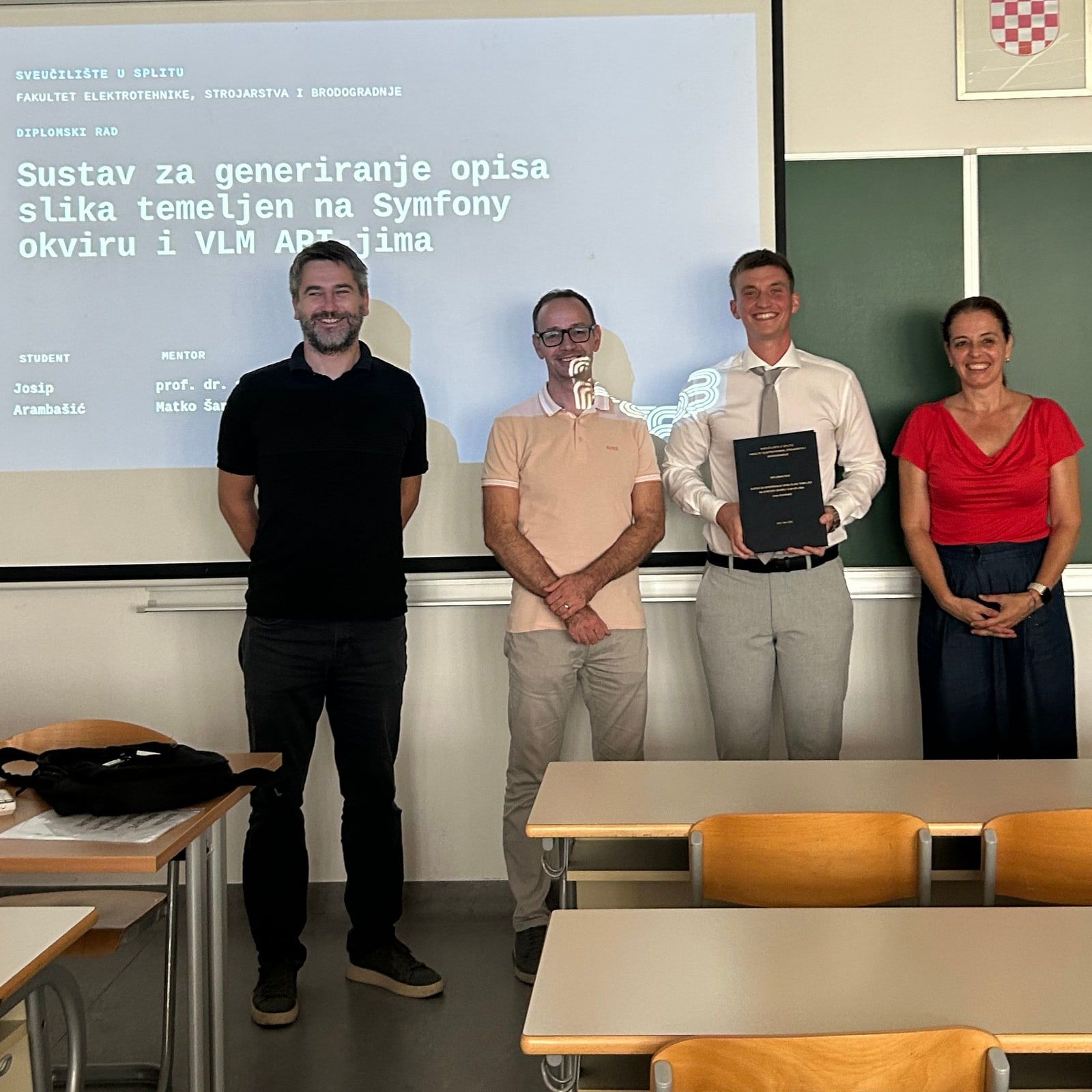 Josip Arambašić holding his bound Master’s thesis, standing with three people in front of the projected title slide at FESB